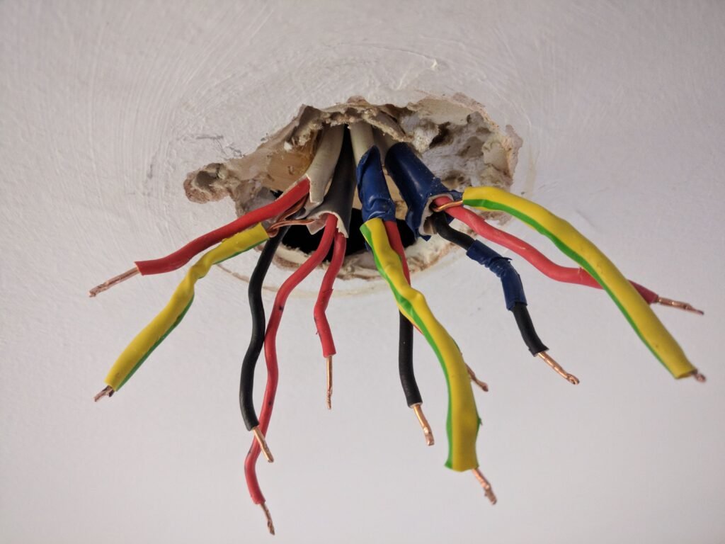 Exposed multicoloured electrical wires protruding from a ceiling, indicative of an in-process light fitting task in the UK, with safety insulating tape on connections.