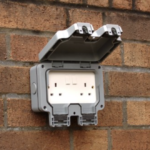 Weatherproof outdoor electrical socket installed on a brick wall, lid open showing the power outlets.