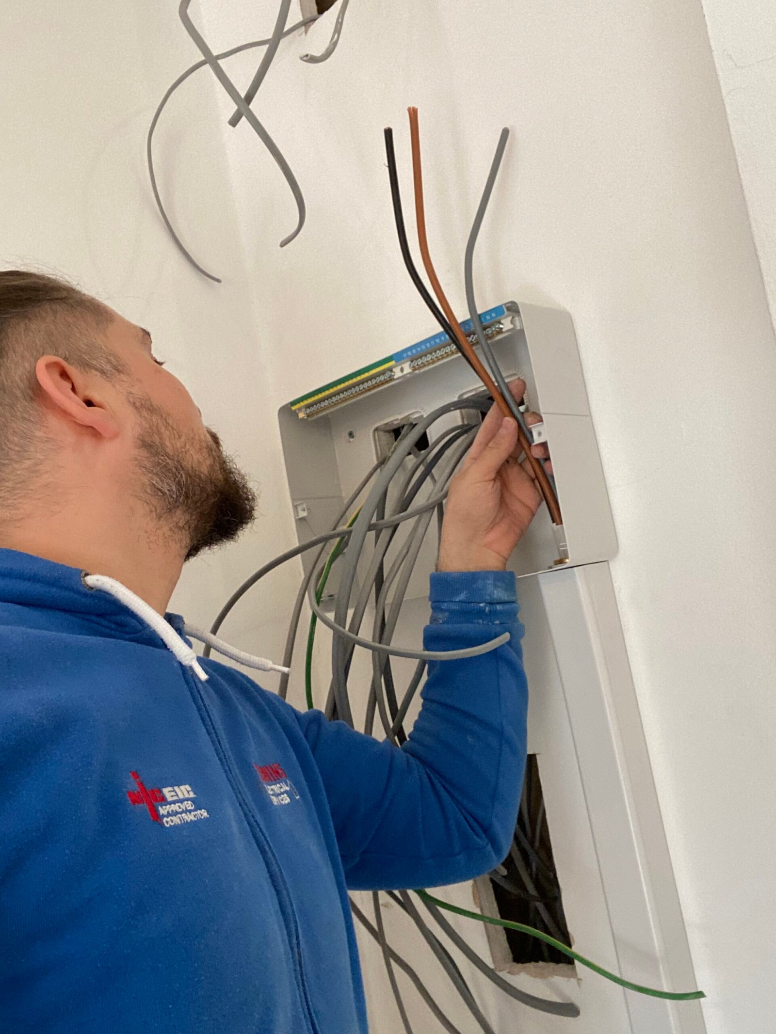 Electrician in blue uniform performing a fuse box rewire, surrounded by multiple cables in East London home.