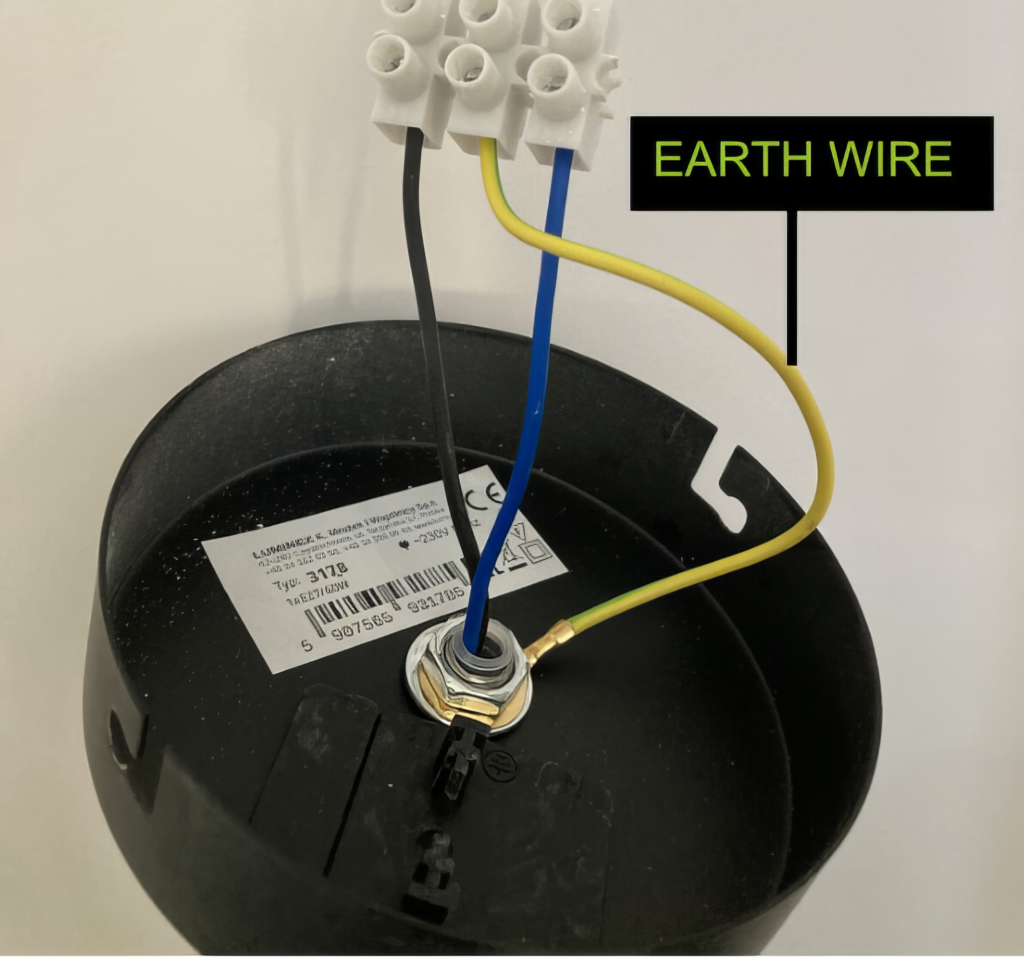 Close-up of an electrical lamp's wiring showing a yellow and green earth wire connected to the lamp base, with a label and terminal block in the background.